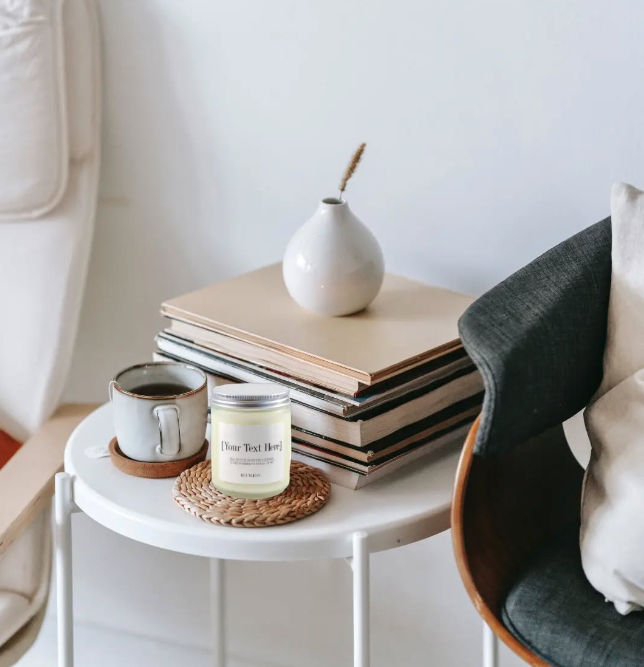 Scandinavian-style side table in a cozy living room with a coffee mug, scented candle on a woven coaster, stacked books and a small white vase.