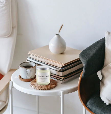 Scandinavian-style side table in a cozy living room with a coffee mug, scented candle on a woven coaster, stacked books and a small white vase.