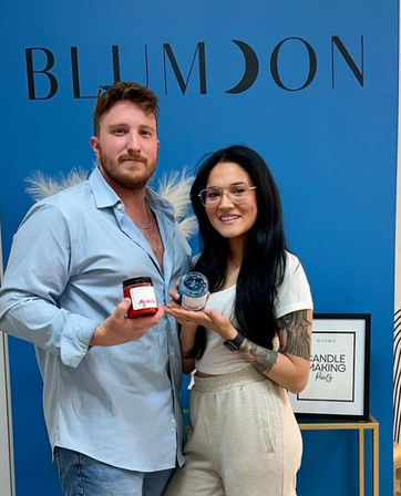 Two people smiling and holding handcrafted candles while posing in a bright blue indoor candle-making workshop.