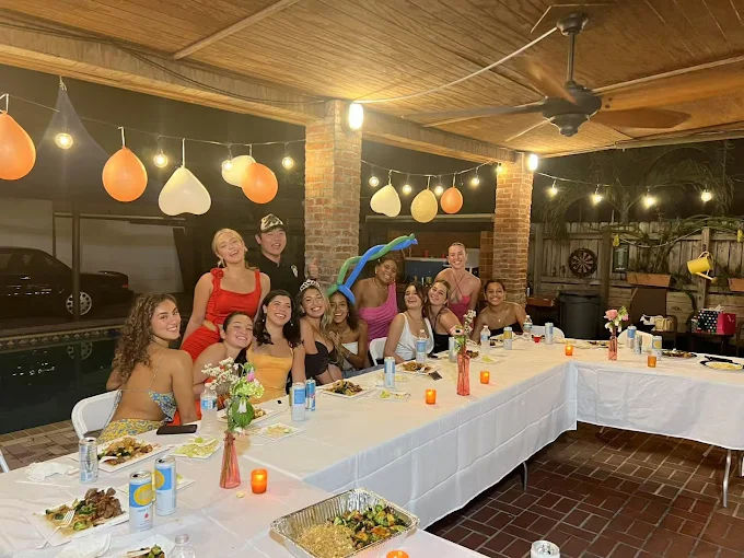 Group of friends at a poolside backyard patio dinner party—long table with food, string lights, balloons, and festive decorations.