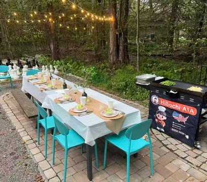 Outdoor garden dinner setup on a stone patio: long table with white tablecloth, turquoise chairs, woven place settings and salads, string lights overhead, portable griddle cooking vegetables beside a propane tank, wooded backdrop and oversized outdoor chess pieces.