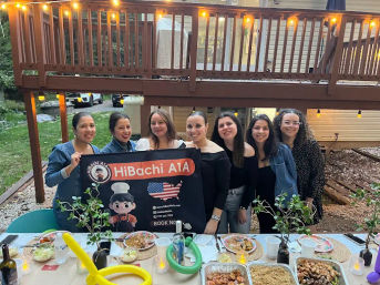 Seven women smiling behind a black promotional banner with a cartoon chef and U.S. flag at a backyard dinner party under string lights, buffet trays and plates on the table.