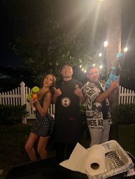Three people posing at a backyard nighttime party under string lights by a white picket fence — woman in a sparkly dress and two men, one in a chef apron, playfully holding colorful water guns.