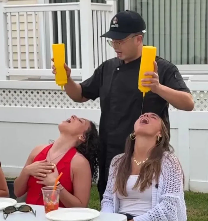 Person in black chef jacket and cap playfully pouring yellow condiment from squeeze bottles into two friends' open mouths at a backyard summer barbecue, white fence, picnic table with plates, sunglasses and a plastic cup visible.
