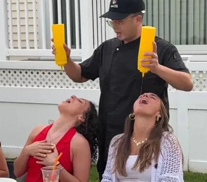 Person in black chef jacket and cap playfully pouring yellow condiment from squeeze bottles into two friends' open mouths at a backyard summer barbecue, white fence, picnic table with plates, sunglasses and a plastic cup visible.