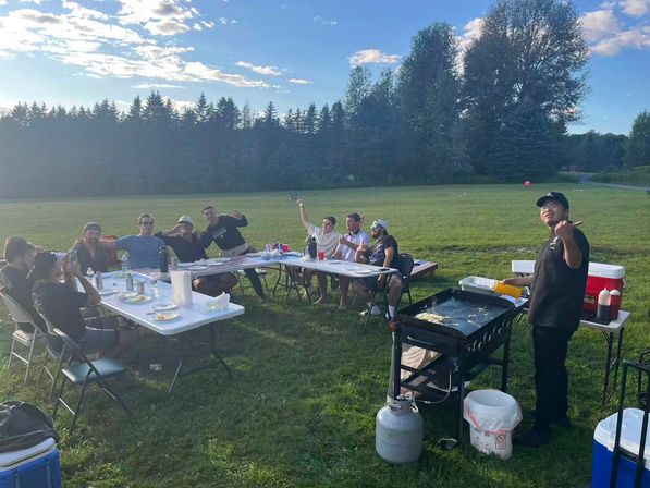 Cheerful summer outdoor picnic in a large grassy field — friends seated at folding tables with plates and drinks, a cook at a portable griddle near coolers, tree line and blue sky.