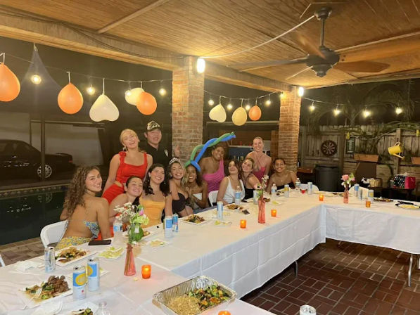 Group of friends at a poolside backyard patio dinner party—long table with food, string lights, balloons, and festive decorations.