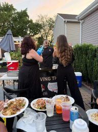 Residential backyard patio at sunset with two women aiming colorful bottles toward a person at an outdoor catering cart, picnic table in foreground with plates and drinks