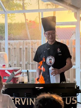 Smiling hibachi chef in a tall hat cooking on a backyard griddle with bright orange flames under a screened patio at sunset