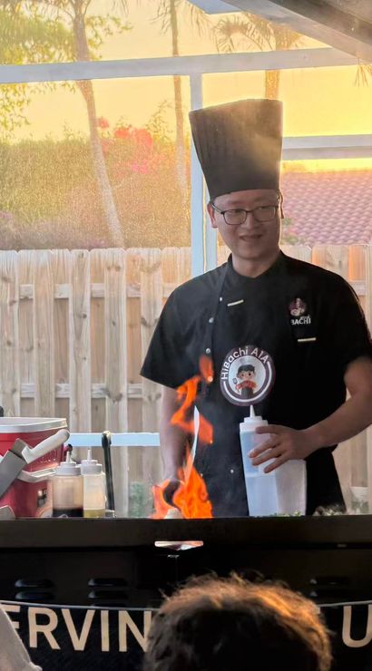 Smiling hibachi chef in a tall hat cooking on a backyard griddle with bright orange flames under a screened patio at sunset