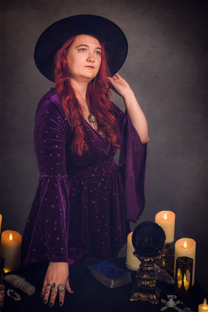 Person with long red hair in a black wide-brim hat and purple celestial velvet dress standing at a table with tarot cards, a black crystal ball on an ornate stand, assorted crystals and glowing candles — mystical tarot setup.