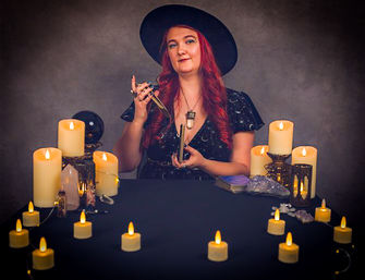 Candlelit tarot-reading portrait of a woman with long red hair and a wide-brim hat holding tarot cards and a pendulum at a table arranged with lit pillar candles, string tealights, crystals, a crystal ball, and a tarot deck.