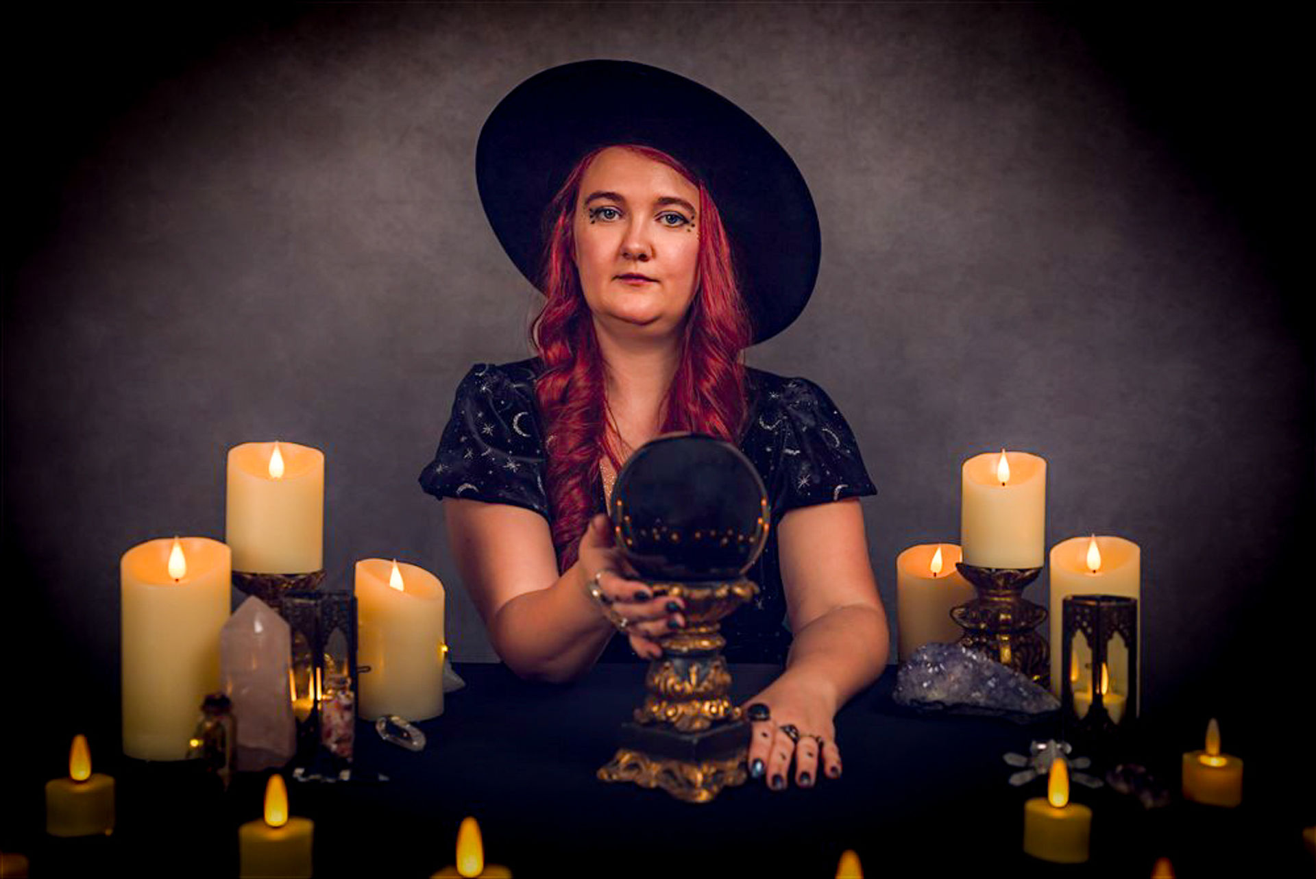 Moody studio portrait of a red-haired fortune-teller in a wide-brim hat holding a crystal ball surrounded by lit candles and crystals