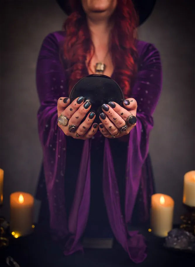Person in a purple robe with red hair holding a black crystal ball above a candlelit altar, tattooed and ringed hands creating a witchy fortune-teller vibe.