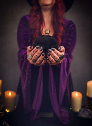 Person in a purple robe with red hair holding a black crystal ball above a candlelit altar, tattooed and ringed hands creating a witchy fortune-teller vibe.