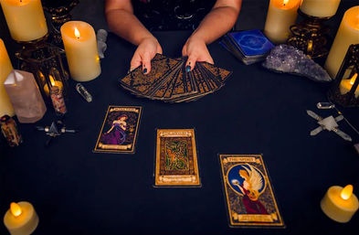 Mystical tarot reading scene — hands fanning an ornate tarot deck on a black cloth, three face-up tarot cards in a spread surrounded by glowing candles, amethyst crystal and scrying stones.