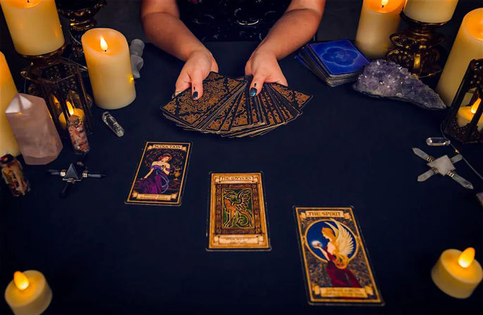 Mystical tarot reading scene — hands fanning an ornate tarot deck on a black cloth, three face-up tarot cards in a spread surrounded by glowing candles, amethyst crystal and scrying stones.