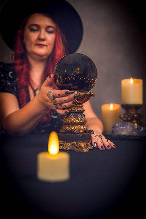 Candlelit psychic scene: woman in a black hat with red hair touches an ornate gold crystal ball on a dark table surrounded by glowing candles.
