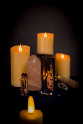 Moody home altar scene with warm glowing candles, a rose quartz crystal tower, small herb-filled glass vials and a black crystal pendulum on dark cloth — cozy spiritual/meditation display.