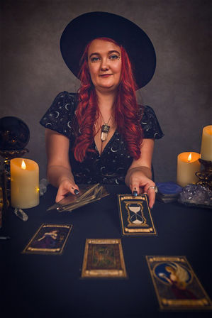 Red-haired tarot reader in a wide black hat and star-patterned dress shuffles tarot cards over a black cloth table with lit candles, a crystal ball and amethyst cluster, several ornate tarot cards laid out including an hourglass card.