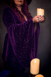 Figure in a purple velvet robe patterned with moons and stars holds a glowing candle above an altar with burning pillar candles and a tarot deck, evoking a mystic tarot reading and spiritual ritual.