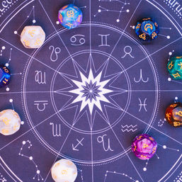 Flat-lay astrology wheel playmat with a white starburst center, zodiac symbols and constellation lines, surrounded by colorful polyhedral dice engraved with zodiac glyphs.