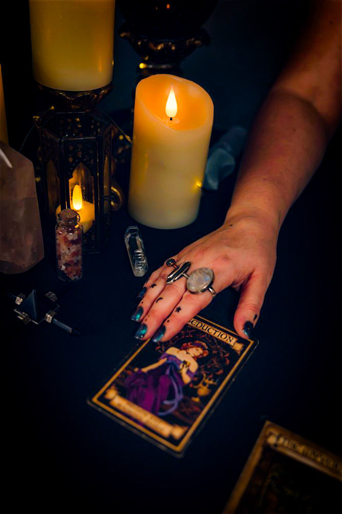 Mystical tarot reading: a ringed hand rests on a 'Seduction' tarot card surrounded by glowing candles, crystal points and a tiny crystal vial on a dark table.