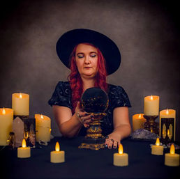 Moody studio portrait of a mystic in a black wide-brim hat and red hair holding a black crystal ball on an ornate gold stand, surrounded by glowing candles and crystals on a dark table.