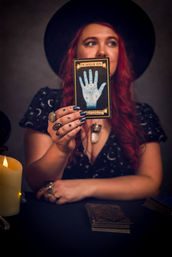 Candlelit tarot reading: a fortune-teller in a wide-brim hat with long red hair holds up a blue palmistry card labeled “The Hand of Fate” above a deck of tarot cards, wearing rings and a crystal pendant in a moody indoor scene.