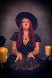 Moody studio portrait of a red-haired tarot reader in a black witch hat and starry dress, holding a fanned deck of tarot cards amid lit candles, crystals, and a crystal pendant necklace — mystical divination scene.