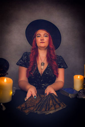 Moody studio portrait of a red-haired tarot reader in a black witch hat and starry dress, holding a fanned deck of tarot cards amid lit candles, crystals, and a crystal pendant necklace — mystical divination scene.