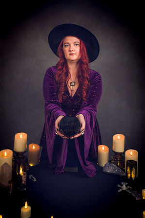 Studio portrait of a fortune-teller in a black wide-brim hat and purple velvet robe holding a black crystal ball above a candlelit altar with amethyst and quartz crystals