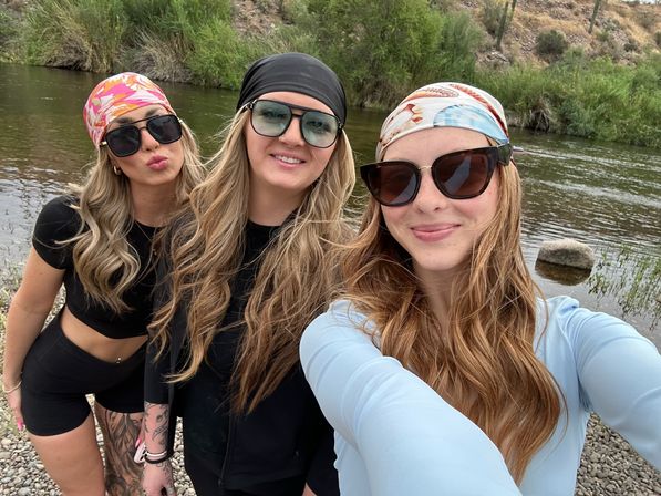Three friends wearing colorful bandanas and sunglasses taking a selfie on a rocky riverbank—one making a kissy face—with a calm river, green shrubs and arid hills in the background.