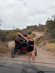 Smiling woman in black activewear posing with raised arm next to a red-and-black off-road UTV on a rocky desert trail with cacti and red rock formations under a cloudy sky