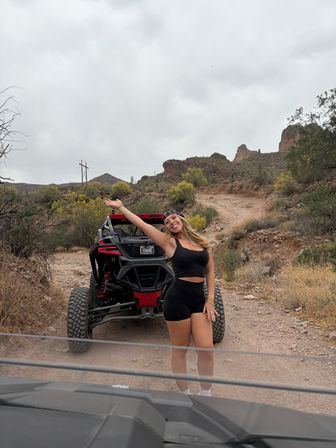 Smiling woman in black activewear posing with raised arm next to a red-and-black off-road UTV on a rocky desert trail with cacti and red rock formations under a cloudy sky