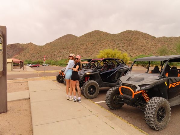 Two women in bandanas posing beside parked off-road UTVs and side-by-sides at a desert trailhead parking lot with scrub-covered hills and a cloudy sky