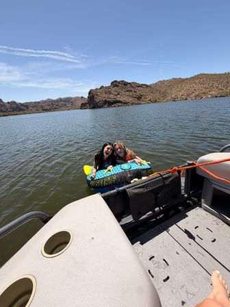 Two friends laughing on a blue towable inflatable behind a boat on a sunny desert lake with rocky hills and a clear blue sky, summer boat tubing scene.