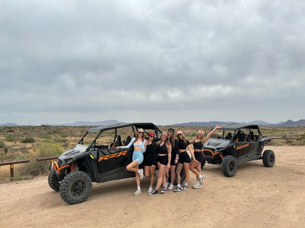 Seven women in athletic outfits posing by two off-road UTV side-by-sides on a dusty desert trail with scrub brush, distant mountains and a cloudy sky