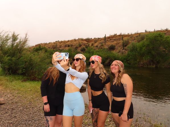 Four friends in activewear and colorful bandanas take a selfie on a rocky desert riverbank with green brush and tall cacti on a dry hillside