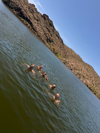 Four swimmers smiling and waving in a green desert lake with rocky, cactus-covered hills and a clear blue sky.