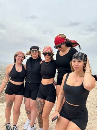 Five women in black activewear and colorful bandanas posing on a windy rocky hilltop overlook above a sprawling city under a cloudy sky after a hike.