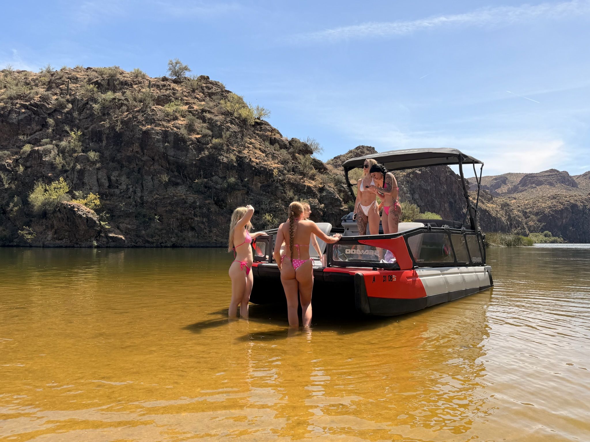 Four women in bikinis on a red pontoon boat in a shallow desert canyon lake with rocky cliffs and a clear blue sky — sunny summer boating scene.
