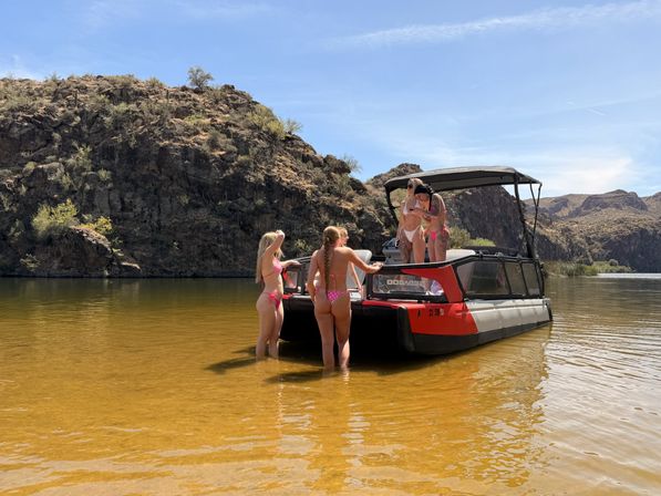 Four women in bikinis on a red pontoon boat in a shallow desert canyon lake with rocky cliffs and a clear blue sky — sunny summer boating scene.