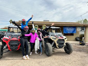 Group of six helmeted riders posing with arms raised between red and tan side-by-side UTVs parked outside a desert powersports rental under an overcast sky
