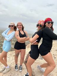 Hiking group of four women in activewear, sunglasses and colorful bandanas posing confidently back-to-back on a rocky hilltop overlooking a sprawling desert valley under a cloudy sky.