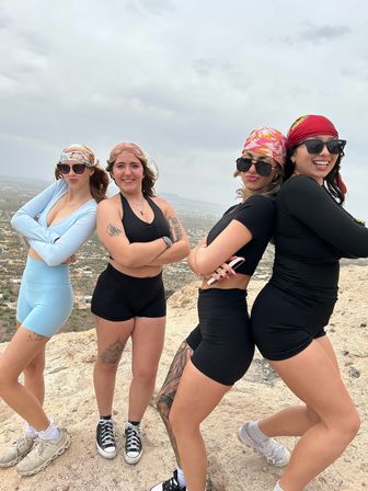 Hiking group of four women in activewear, sunglasses and colorful bandanas posing confidently back-to-back on a rocky hilltop overlooking a sprawling desert valley under a cloudy sky.