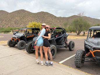 Two women wearing bandanas and sunglasses pose back-to-back by orange-accented off-road UTVs in a desert parking lot with scrubby trees and rocky foothills under a cloudy sky.
