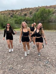 Four women in black activewear playfully sticking out their tongues while walking on a rocky desert riverbank with cacti, scrub brush and arid hills under an overcast sky