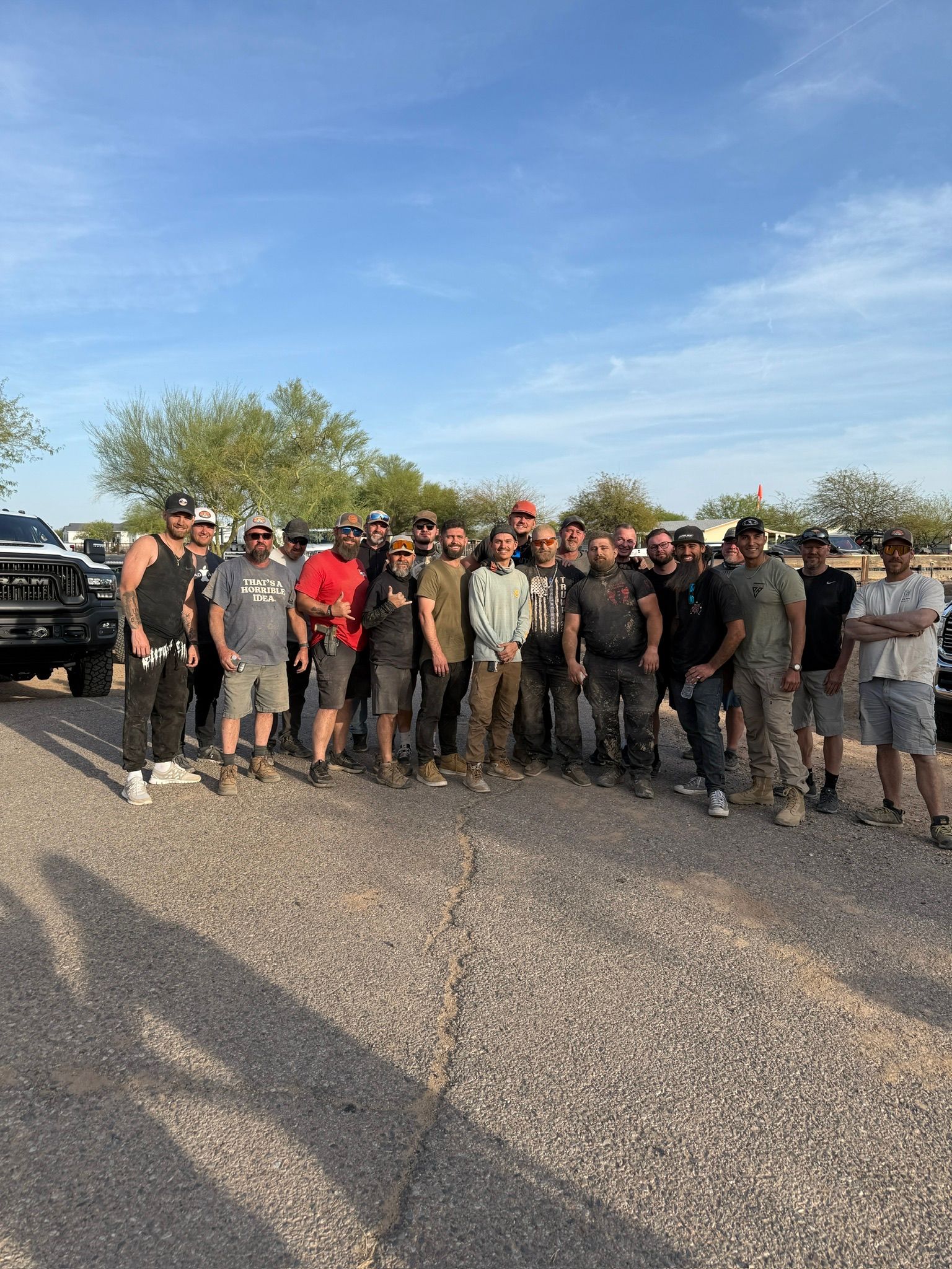 Around 20 men in casual, dusty work clothes posing together in a sunny desert parking lot with pickup trucks and low scrub trees under a bright blue sky.
