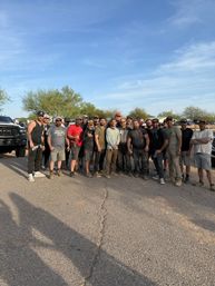 Around 20 men in casual, dusty work clothes posing together in a sunny desert parking lot with pickup trucks and low scrub trees under a bright blue sky.